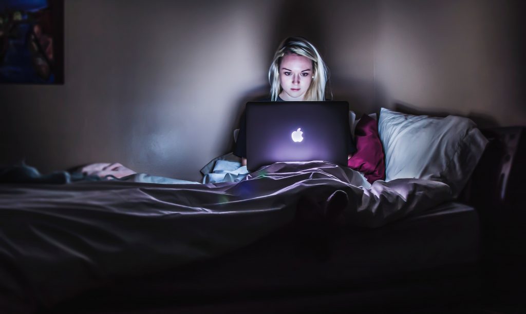 Nainen istuu pimeässä sängyllä tietokoneen ruutu auki ja valo heijastuu ruudulta hänen kasvoilleen.