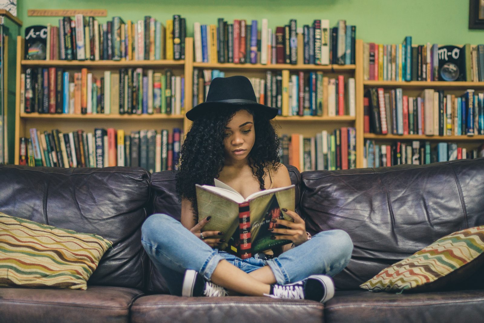 Nainen istuu kirjahyllyn edessä sohvalla ja lukee kirjaa