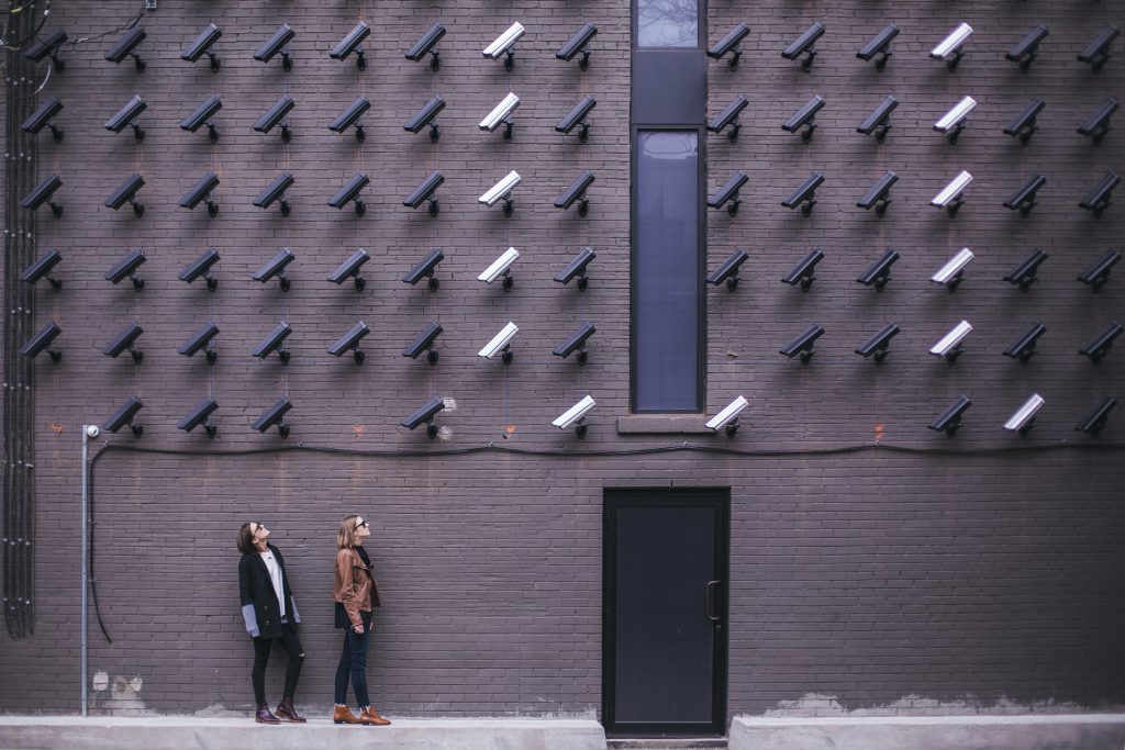 Kaksi ihmistä seisoo talon seinän edessä, missä on koko seinän verran valvontakameroita kohdistettuna heihin.