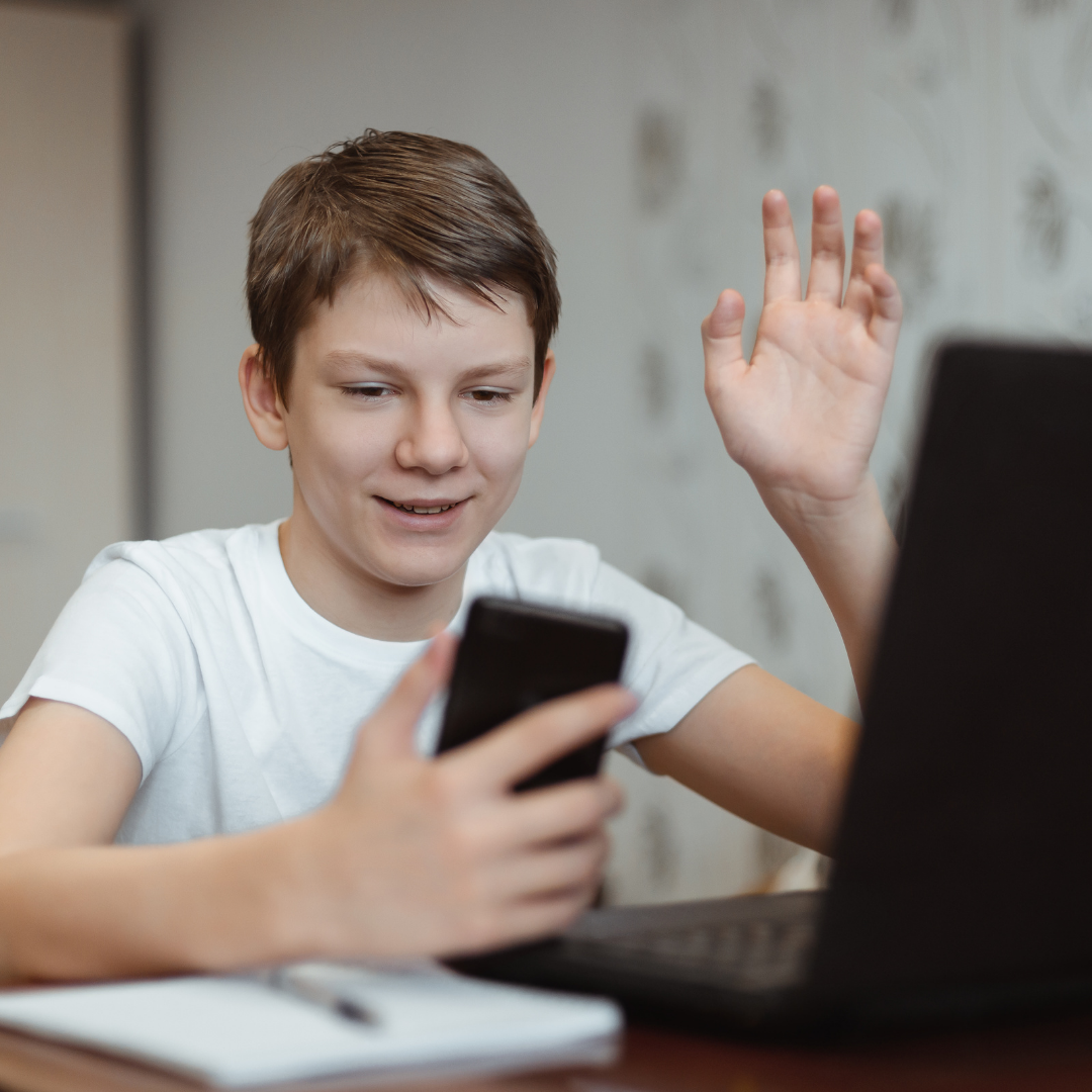 Poika istuu tietokoneen edessä ja heiluttaa kättä kädessä olevaan puhelimeen.