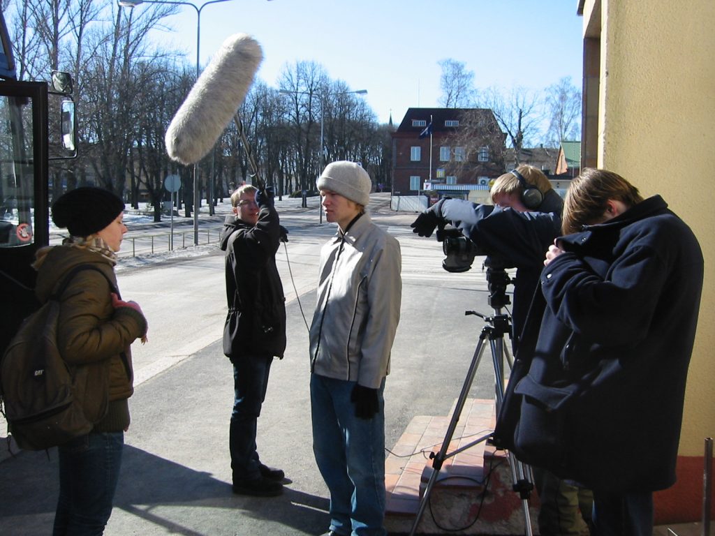 Kadulla kuvataan videota. Yksi henkilö kuvaa videokameralla, yksi pitelee mikkiä ja kaksi henkilöä näyttelee kadulla.